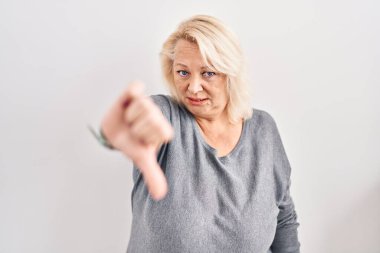 Middle age caucasian woman standing over white background looking unhappy and angry showing rejection and negative with thumbs down gesture. bad expression. 