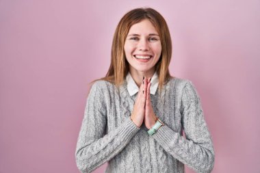Beautiful woman standing over pink background praying with hands together asking for forgiveness smiling confident. 