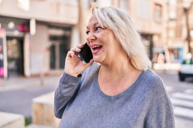 Orta yaşlı, sarışın, gülümseyen, kendine güvenen bir kadın caddede akıllı telefondan konuşuyor.
