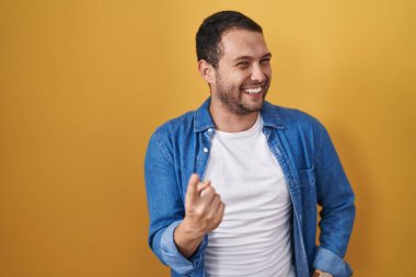 Hispanic man standing over yellow background beckoning come here gesture with hand inviting welcoming happy and smiling 