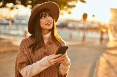 Kışlık şapkalı esmer kadın gün batımında şehrin dışında akıllı telefon kullanarak gülümsüyor.
