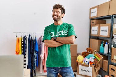 Young hispanic volunteer man smiling happy standing with arms crossed gesture at charity center.