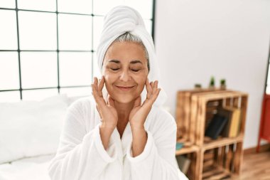 Yatak odasındaki yatakta oturan, yüz kremi kullanan orta yaşlı, kır saçlı bir kadın..