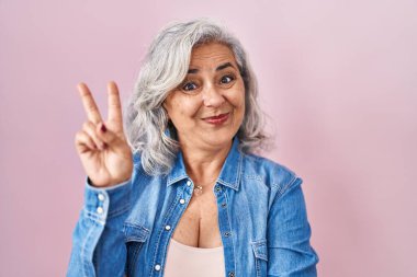 Pembe arka planda duran gri saçlı orta yaşlı kadın kameraya gülümseyen ve parmaklarıyla zafer işareti yapan. İki numara.. 