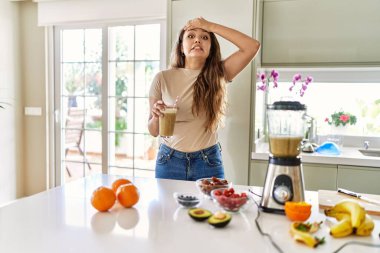 Güzel, genç, esmer bir kadın mutfakta bir bardak smoothie içiyor. Stresli ve hayal kırıklığına uğramış, şaşırmış ve kızgın bir yüzü var. 
