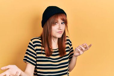 Kızıl saçlı, yünlü şapka takan, elleri ve kolları havada, kafası karışmış genç bir kadın. şüphe kavramı. 