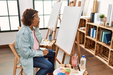 Orta yaşlı İspanyol bir kadın resim stüdyosunda tuvale resim yapıyor ağzı elle kaplıyor, şok oluyor ve hata yapmaktan korkuyor. şaşırmış ifade 