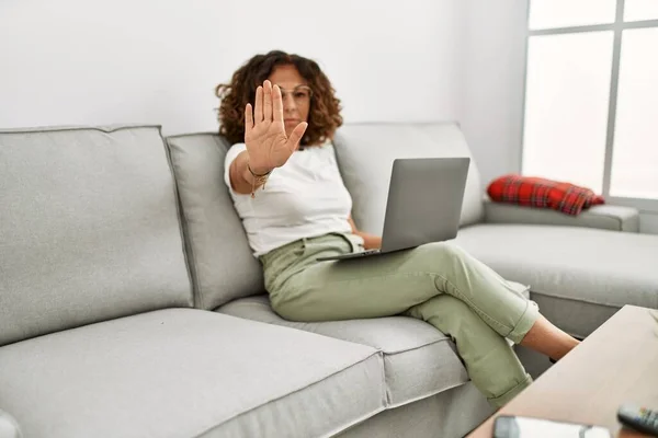 Middle age hispanic woman working using computer laptop at home with open hand doing stop sign with serious and confident expression, defense gesture