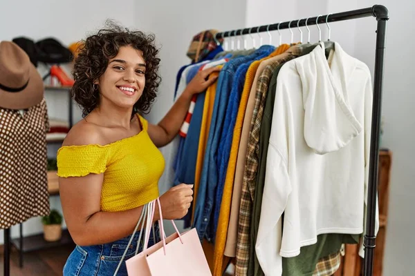 Young latin customer woman smiling happy choosing clothes and holding shopping bags at clothing store.