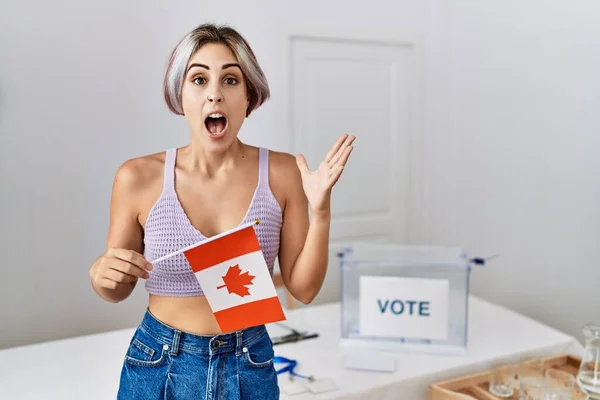 Young beautiful woman at political campaign election holding canada flag celebrating victory with happy smile and winner expression with raised hands