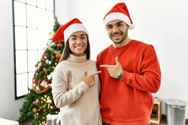笑顔で陽気にクリスマスツリーのそばに立っている若いヒスパニック系のカップルは 幸せと自然な表情で側に手と指を指しています — ストック写真
