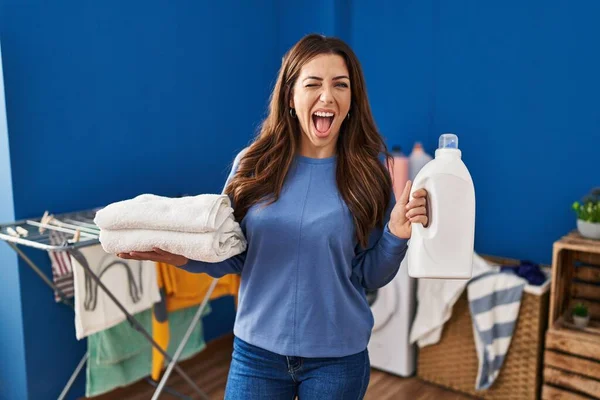 Young brunette woman holding clean laundry and detergent bottle winking looking at the camera with sexy expression, cheerful and happy face.