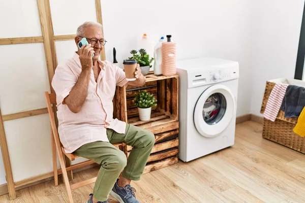 Senior man talking on the smartphone and drinking coffee waiting for washing machine at laundry room