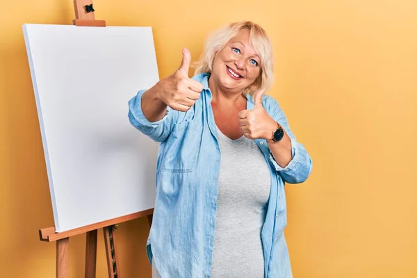 Middle Age Blonde Woman Standing Painter Easel Stand Approving Doing — Stockfoto