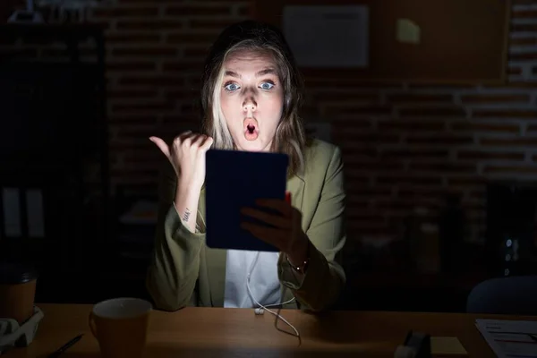 Blonde caucasian woman working at the office at night surprised pointing with hand finger to the side, open mouth amazed expression.