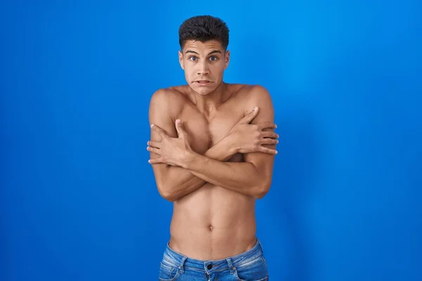 Young hispanic man standing shirtless over blue background shaking and freezing for winter cold with sad and shock expression on face