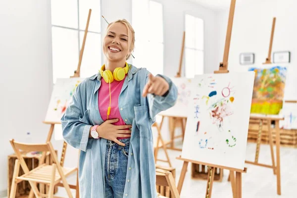 Young caucasian girl at art studio laughing at you, pointing finger to the camera with hand over body, shame expression