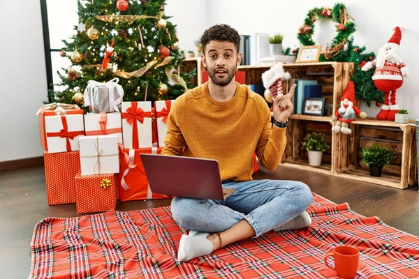 カメラを見ている側に手の指で指して 顔に大きな笑顔でクリスマスツリーに座っているラップトップを使用してアラブの若い男 — ストック写真