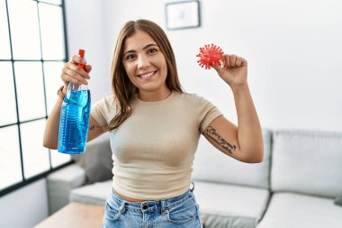 Genç İspanyol kadın, elinde virüs oyuncağı ve dezenfekte spreyi tutarak gülümsüyor.