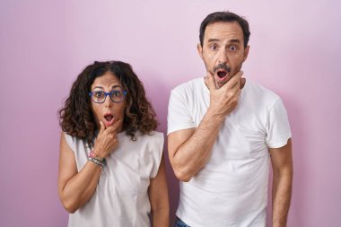 Middle age hispanic couple together over pink background looking fascinated with disbelief, surprise and amazed expression with hands on chin 