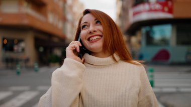Genç kızıl saçlı, gülümseyen, kendine güvenen, caddedeki akıllı telefondan konuşan kadın.