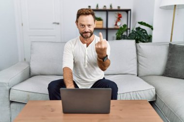 Middle age man using laptop at home showing middle finger, impolite and rude fuck off expression 