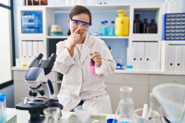 Hispanic girl with down syndrome working at scientist laboratory smelling something stinky and disgusting, intolerable smell, holding breath with fingers on nose. bad smell 