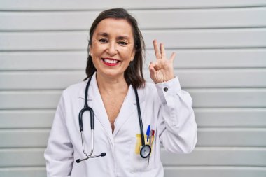 Middle age hispanic woman wearing doctor uniform and stethoscope doing ok sign with fingers, smiling friendly gesturing excellent symbol 