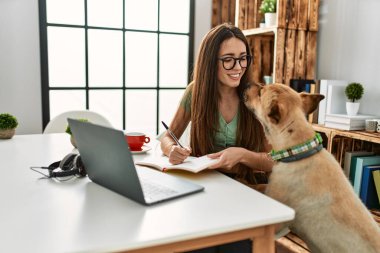 Genç İspanyol kadın dizüstü bilgisayar kullanıyor ve kitap yazıyor. Evde köpekle birlikte masada oturuyor.