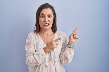 Middle age hispanic woman standing over blue background pointing aside worried and nervous with both hands, concerned and surprised expression 