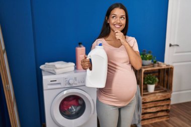 Çamaşır deterjanı şişesini tutan hamile bir kadın. Ciddi bir yüz ifadesi. Çene üstüne çene sorusu, kafa karıştırıcı bir fikir. 