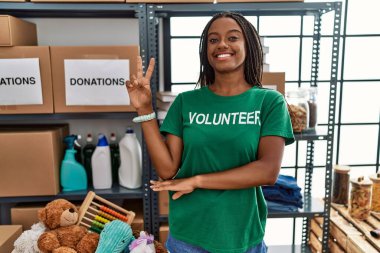 Young african american woman working wearing volunteer t shirt at donations stand smiling with happy face winking at the camera doing victory sign with fingers. number two. 