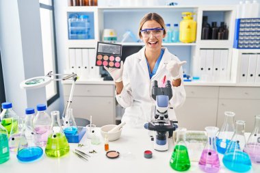 Laboratuvarda çalışan genç sarışın kadın makyajlı ve ağzı açık gülümseyen bir şekilde başparmağını yana kaldırıyor. 