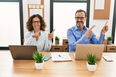 Orta yaşlı İspanyol kadın ve erkek ofiste dizüstü bilgisayarla oturuyor gülümsüyor ve kameraya bakıyor iki eliyle ve parmaklarıyla kenarı işaret ediyor.. 