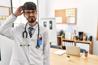 Hispanic man with beard wearing doctor uniform and stethoscope at the office confuse and wonder about question. uncertain with doubt, thinking with hand on head. pensive concept. 