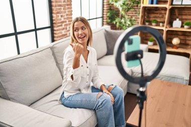 Young blonde woman recording vlog tutorial with smartphone at home pointing thumb up to the side smiling happy with open mouth 