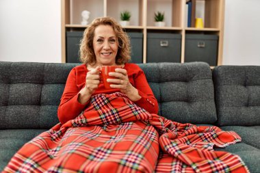 Orta yaşlı beyaz kadın evdeki kanepede kahve içiyor..