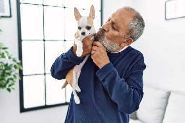 Yaşlı, kır saçlı, gülümseyen, özgüvenli bir adam evde Chihuahua öpüyor.