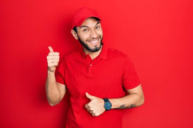 Hispanic man with beard wearing delivery uniform and cap pointing to the back behind with hand and thumbs up, smiling confident 