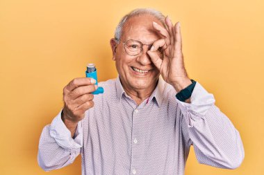 Senior man with grey hair holding medical asthma inhaler smiling happy doing ok sign with hand on eye looking through fingers 