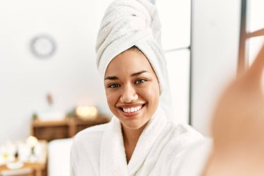 Bornoz giyen genç Latin kadın güzellik merkezindeki kameranın önünde selfie çekiyor.