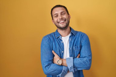 Hispanic man standing over yellow background happy face smiling with crossed arms looking at the camera. positive person. 