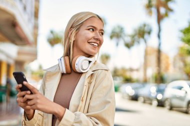 Şehirde akıllı telefon ve kulaklık kullanarak mutlu gülümseyen genç sarışın kız..