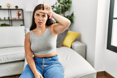 Genç esmer kadın evdeki kanepede oturuyor. Alnında parmakları olan insanlarla dalga geçiyor. Ezik hareketler yapıyor.. 