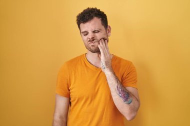 Sarı arka planda duran İspanyol genç adam diş ağrısı ya da diş ağrısı yüzünden acı dolu bir ifadeyle ağıza dokunuyor. dişçi 