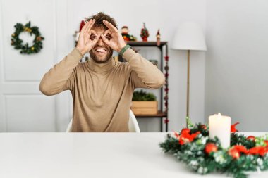 Noel süslemesi sırasında masanın üzerinde oturan sakallı yakışıklı genç adam dürbün gibi jest yapıyor, gözleri parmaklarından bakıyor. çılgın ifade. 