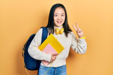 Öğrenci çantası tutan genç Çinli kız ve gülümseyen kitaplar kameraya bakıp zafer işareti yapıyor. İki numara.. 