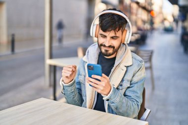 Young hispanic man listening to music sitting on table at coffee shop terrace