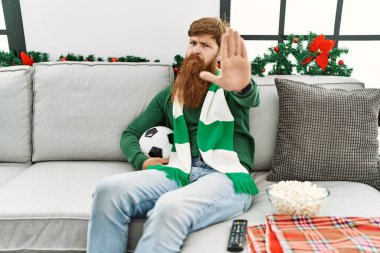 Redhead man with long beard football hooligan holding ball sitting on the sofa with open hand doing stop sign with serious and confident expression, defense gesture 