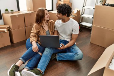 Genç çift yeni evinde dizüstü bilgisayar kullanarak mutlu bir şekilde gülümsüyor..